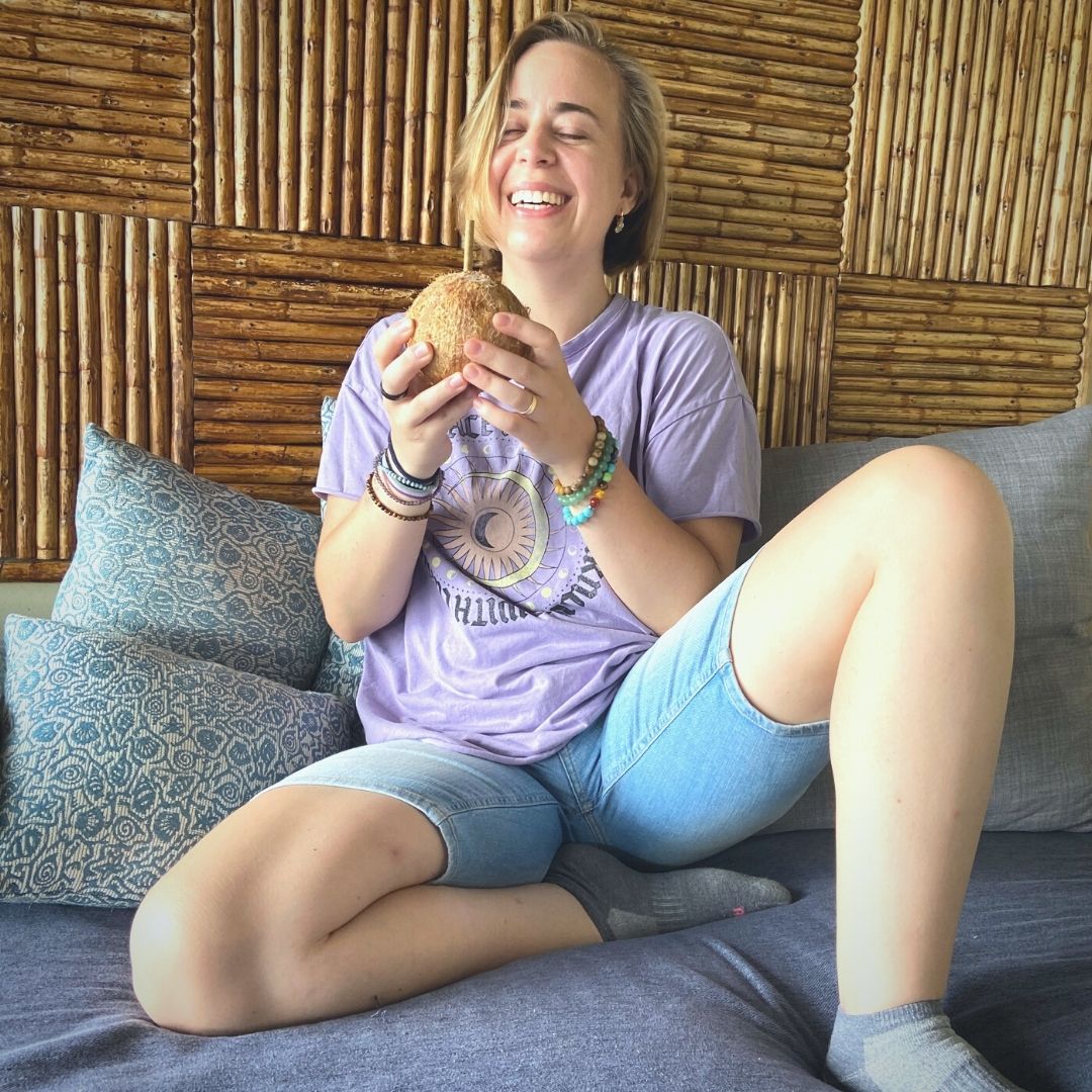 A photo of Sarah holding a coconut while sitting in a cushy bed, laughing to herself and feeling joyful as one does when being asked to pose for pictures.