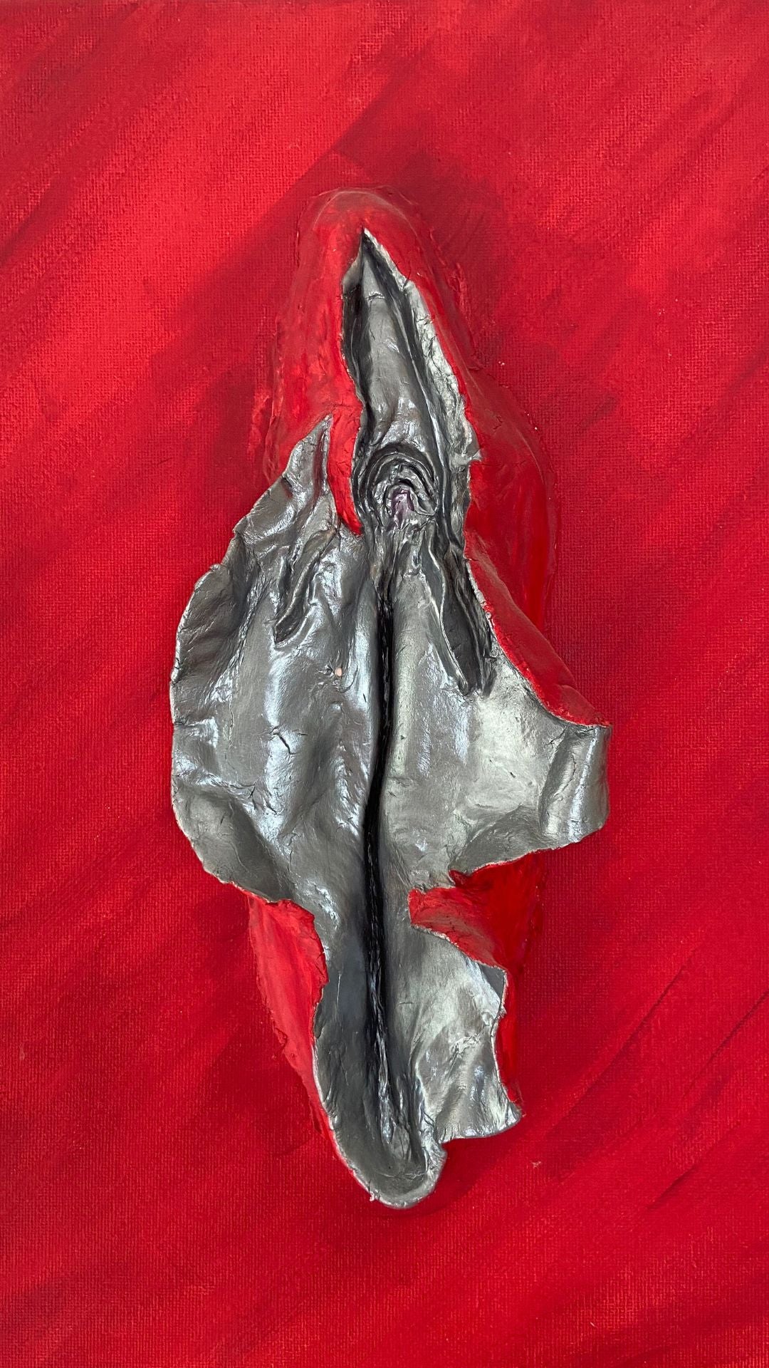 A photo of Sarah Lemmerman's original silver vulva sculpture mounted on red canvas. The inner labia glistens in metallic silver in contrast to the bright red surroundings.