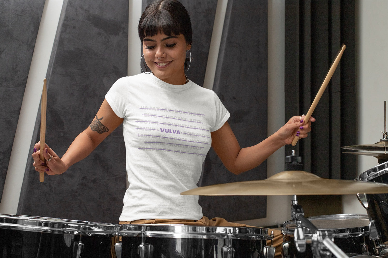 A photo of a person wearing the "Call Me Vulva" t-shirt design while playing the drums with a joyful expression.