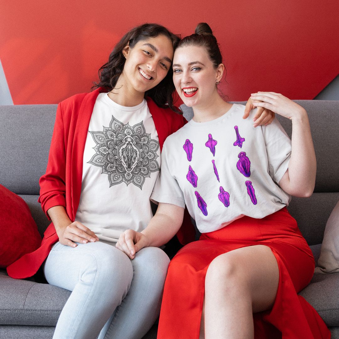 A photo of a two individuals wearing vulva t-shirts while smiling together on a grey couch against a red background.