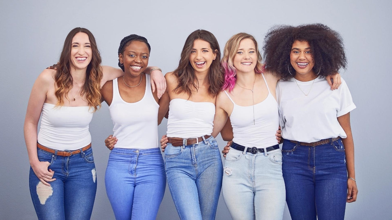 A photo of a jubilant group of five people wearing similar outfits of blue jeans and white t-shirts or tank tops. They have their arms around one another and are smile-laughing while facing you.