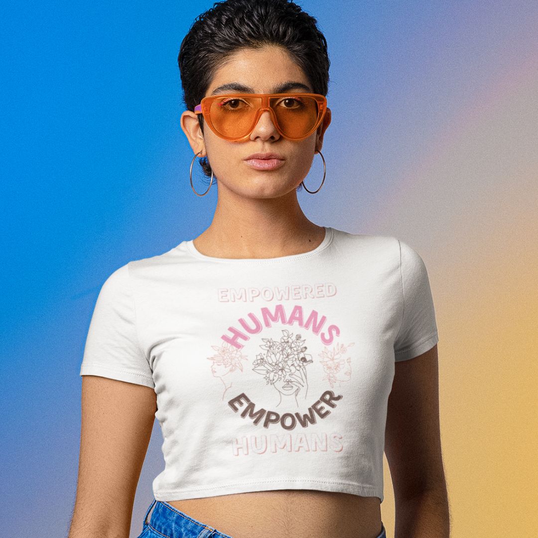Photo of a person wearing orange glasses and a crop top with the "Empowered Humans Empower Humans" design, gazing confidently at you while standing against a blue and orange background.