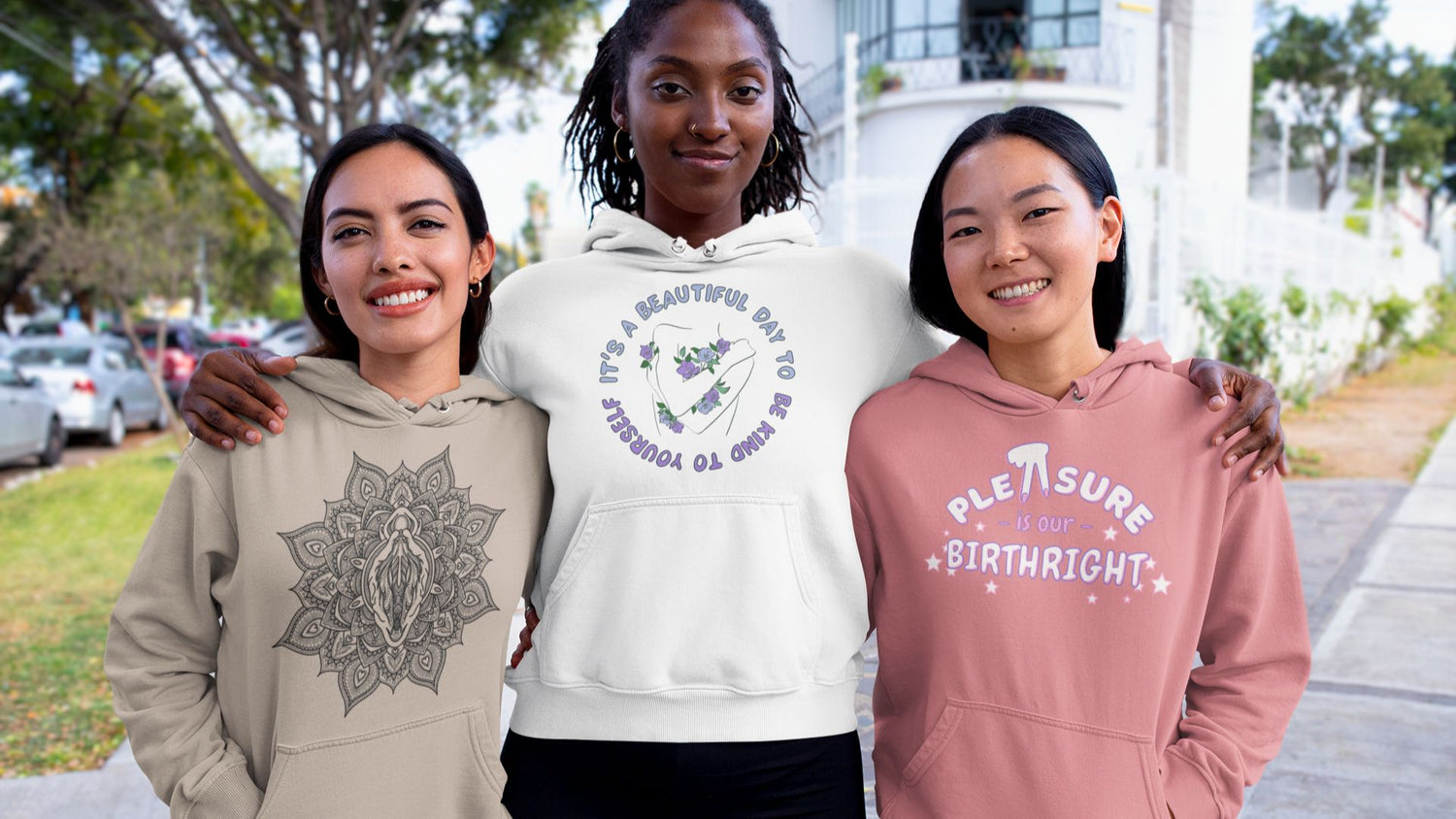 Photo of three people wearing vulva hoodie designs, including from left to right, the vulva mandala, it's a beautiful day to be kind to yourself, and pleasure is our birthright. They're smiling with arms around one another, standing at a bright neighborhood corner.
